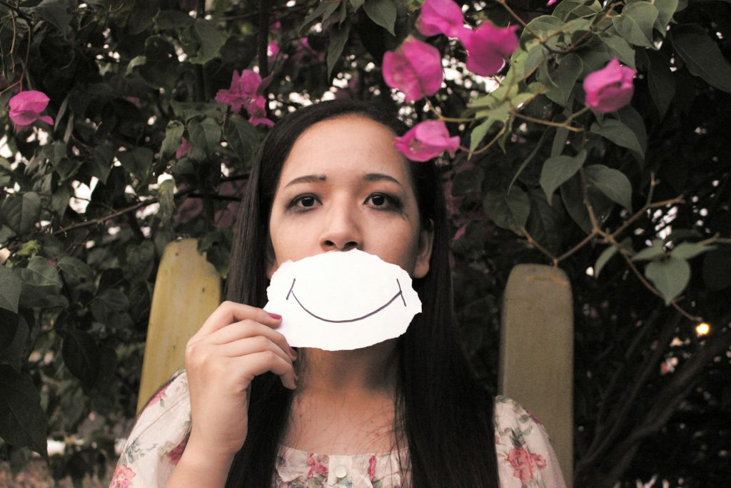 A woman holding a paper with a drawn smile in front of her mouth, surrounded by vibrant pink flowers and greenery, symbolizing hidden emotions.