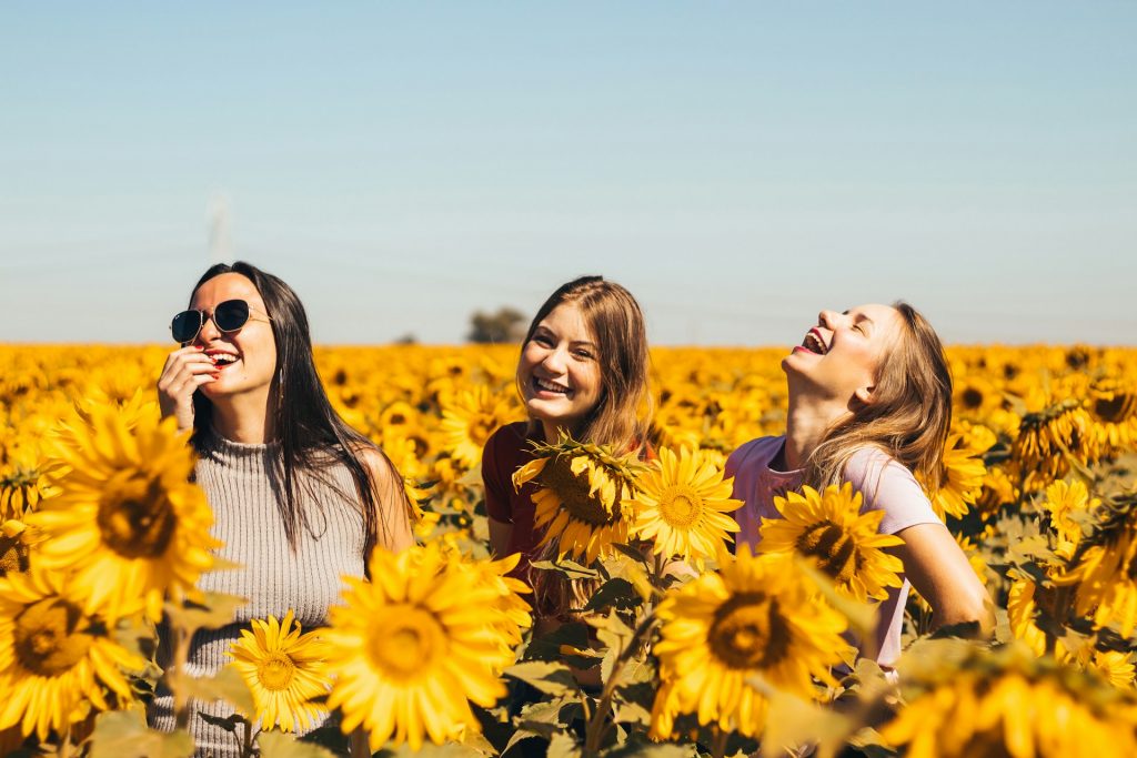 Three women laughing and enjoying a sunny day in a field of vibrant sunflowers, symbolizing joy, connection, and shared experiences.