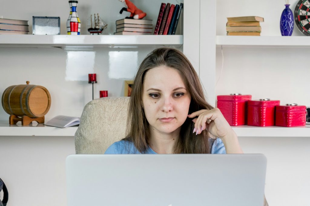 A woman focused on her laptop, seated in a cozy home office with shelves of books and decorative items in the background.