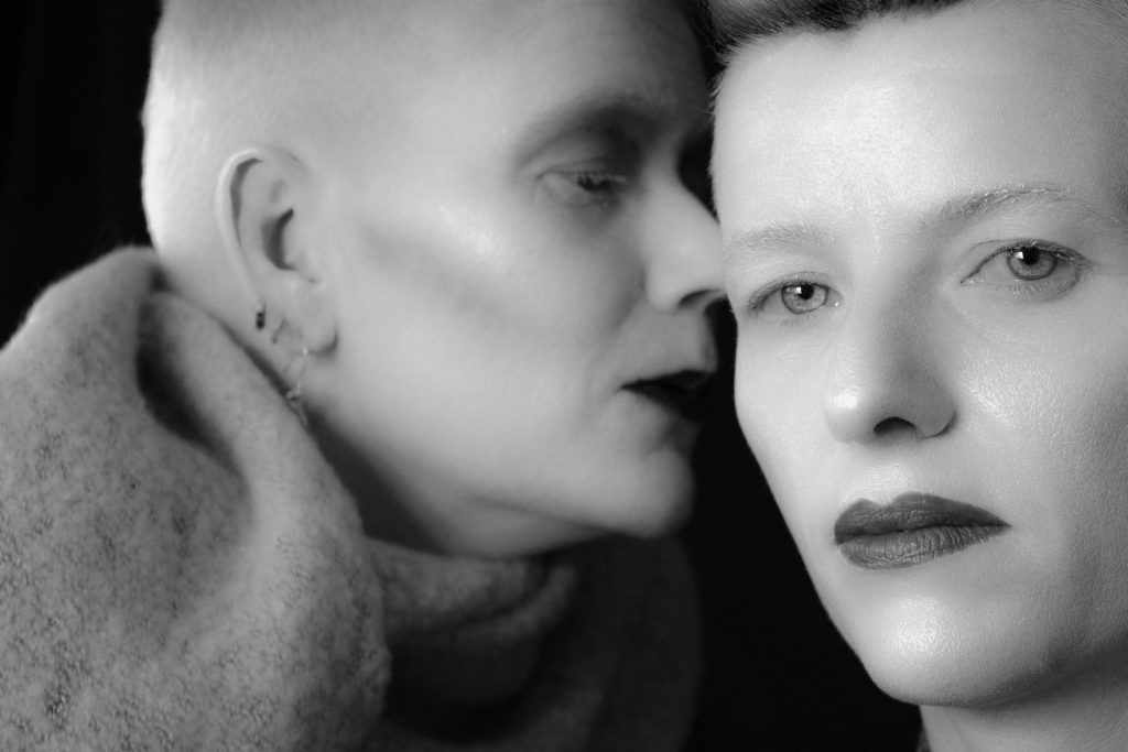 A black-and-white portrait of two individuals. One is whispering into the ear of the other, who gazes directly at the camera with an enigmatic expression.