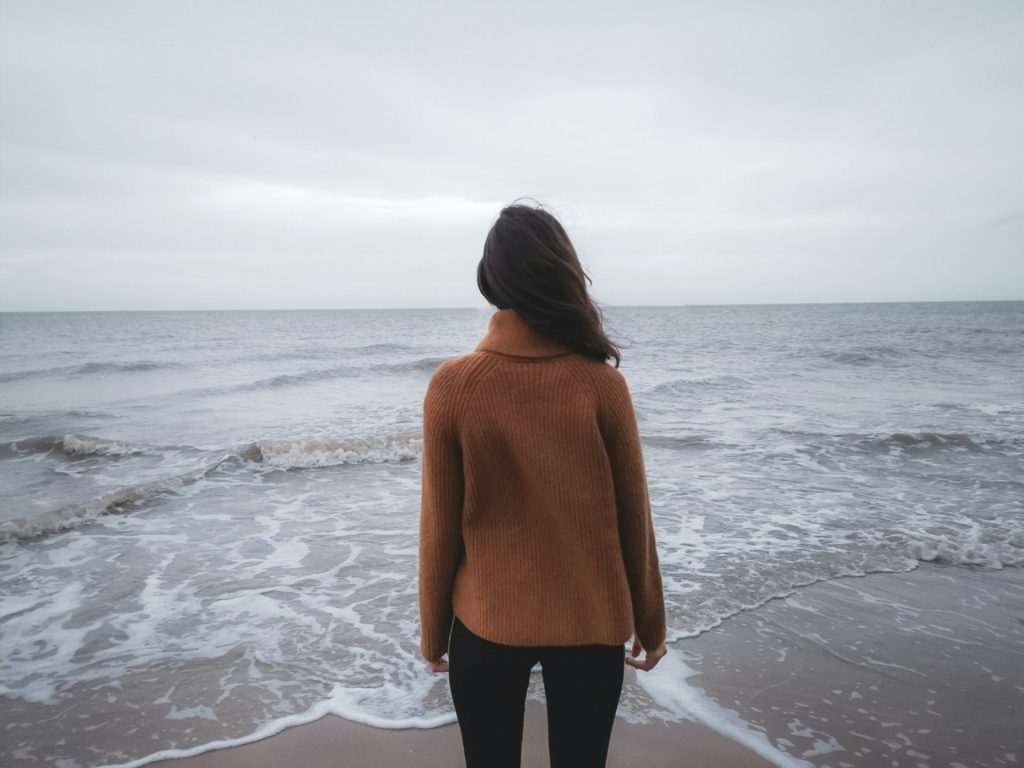 A woman standing on a beach, facing the ocean under a cloudy sky, symbolizing solitude, introspection, and personal reflection.