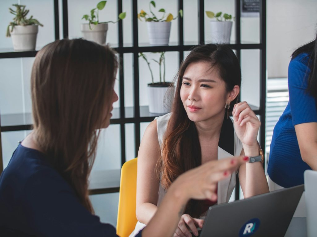 Two women engage in a thoughtful discussion in a modern workspace, one listening intently while the other gestures expressively, conveying collaboration and understanding.