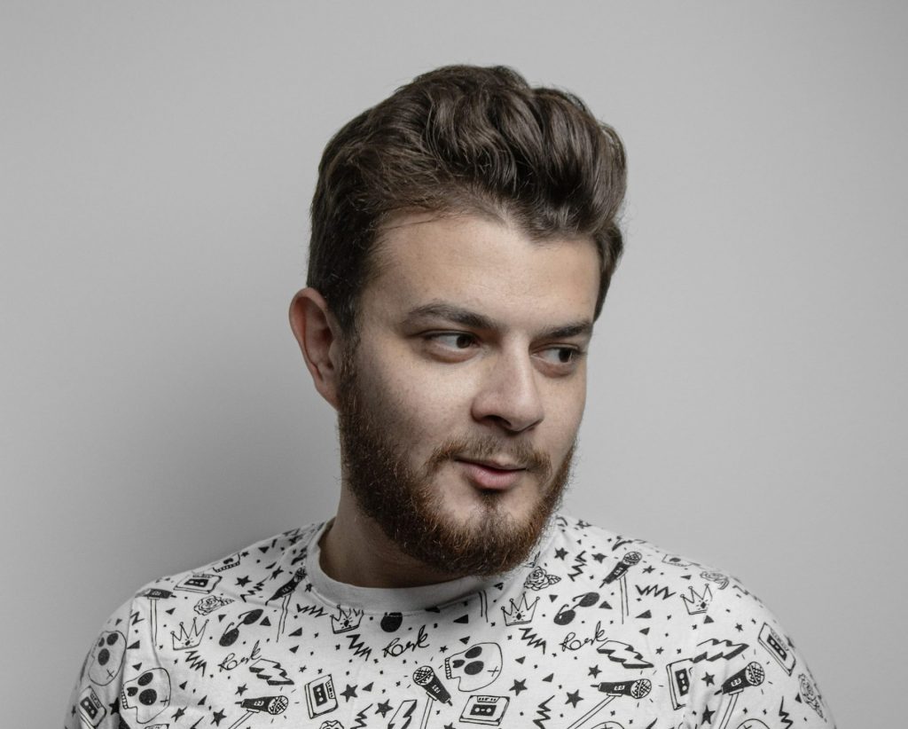 Young man in a printed t-shirt making expressive hand gestures while speaking, standing against a plain gray background.