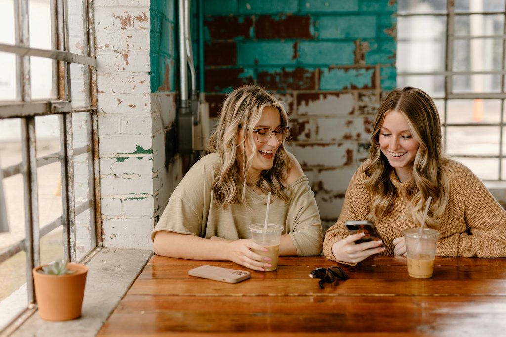 Two women sitting at a wooden table in a rustic café, laughing while looking at a smartphone, with iced coffee and a succulent plant nearby.