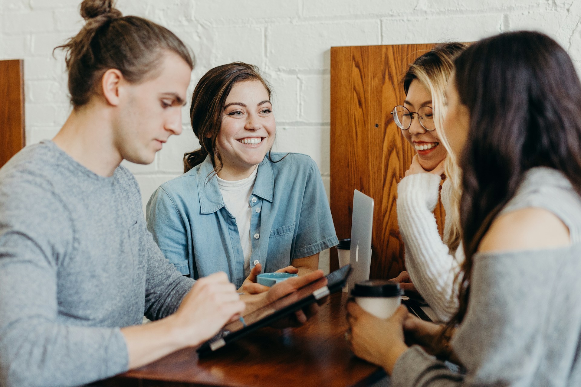 Young adults smiling and talking at a cafe table, working on a tablet and laptop while holding coffee cups.