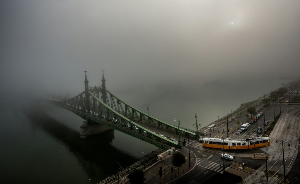 Liberty Bridge in Budapest covered in fog with a yellow tram on the riverbank and cars crossing the bridge.