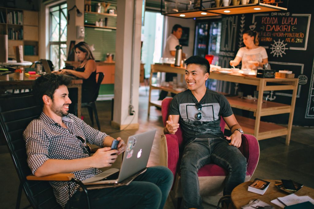 Two men sitting in a café, smiling and talking while working on a laptop, with people and a chalkboard menu visible in the background.