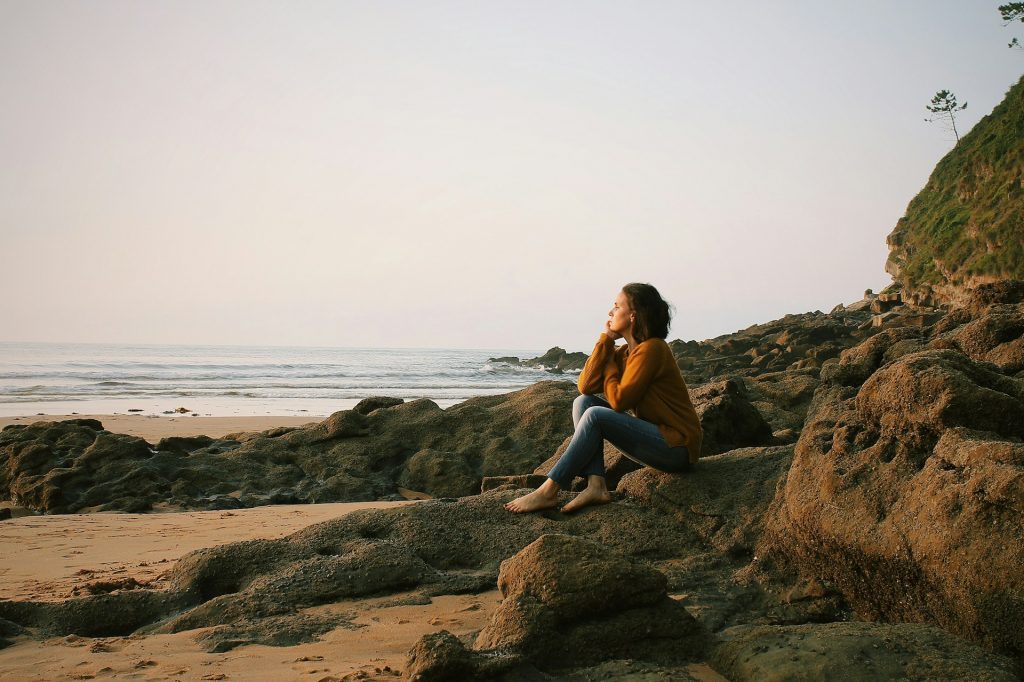 A woman in a mustard sweater sitting on rocks by the ocean, gazing at the horizon under a soft sunrise sky.