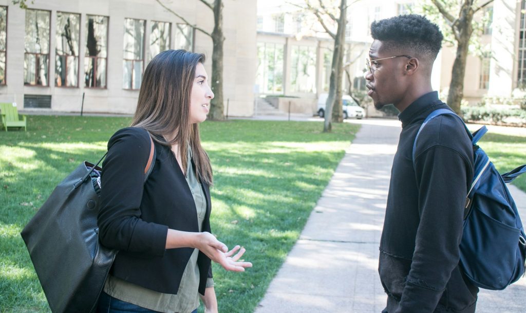 Two students having a conversation outside on a college campus, with green grass and a modern building in the background.
